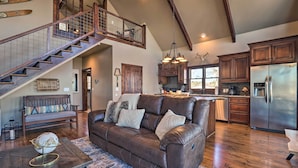 A spacious living area with a brown leather sofa, wooden staircase, and a kitchen with stainless steel appliances. - Shoreline Home w/ Boat Dock & Fire Pit in Shelby! (Shelby)