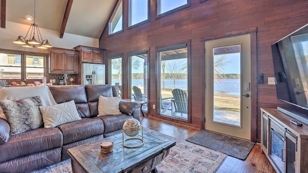 A spacious living room with a leather sofa, a coffee table, and a large window overlooking a body of water. - Shoreline Home w/ Boat Dock & Fire Pit in Shelby! (Shelby)