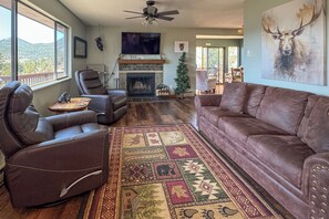 A living room with a fireplace, leather sofas, a large TV, and a mountain view. - 3BR Cabin Near Rmnp Sweeping Views + Fireplace (Estes Park)