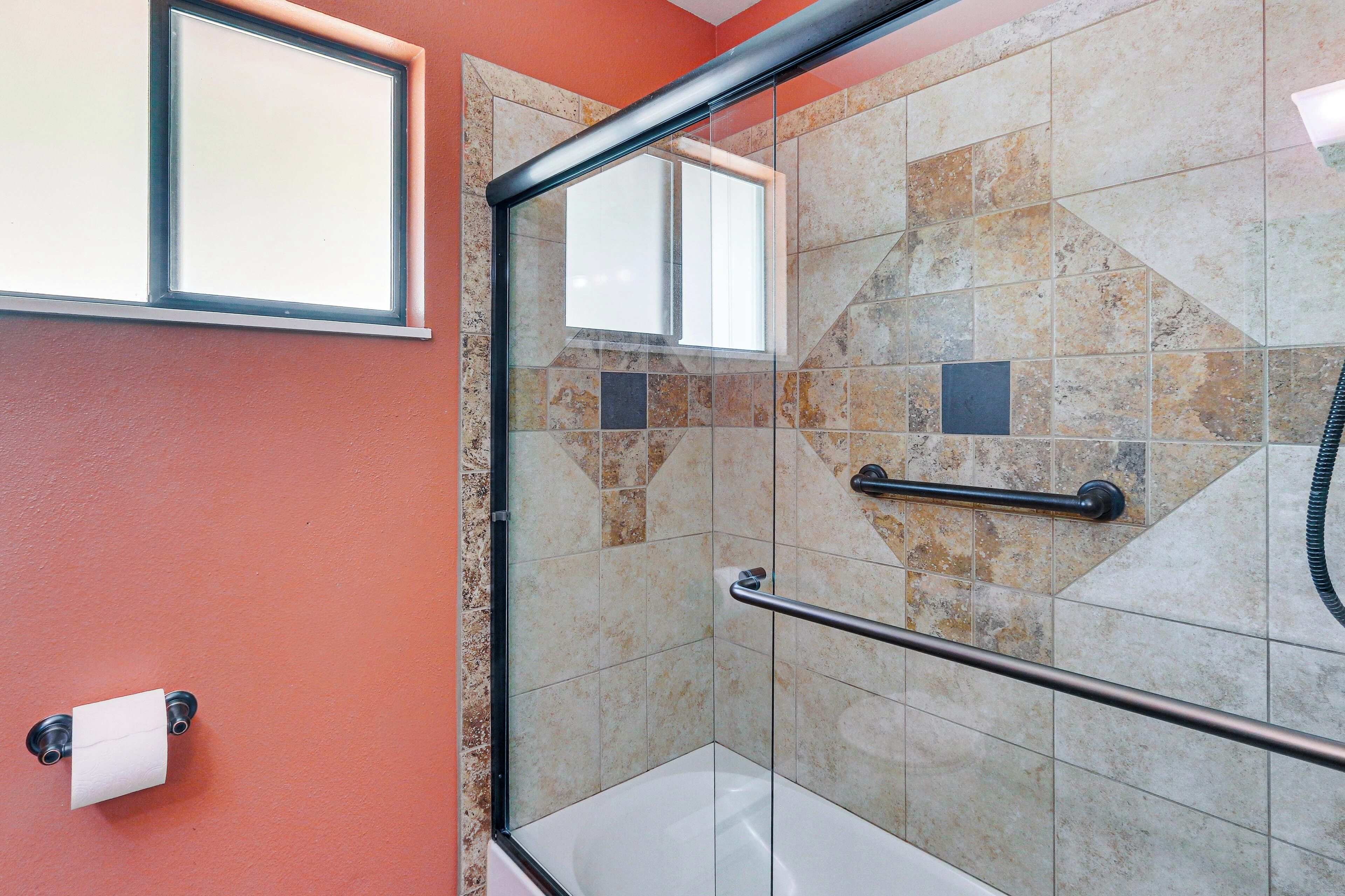 A bathroom with a glass shower enclosure, a handrail, a towel dispenser, and a mirror.