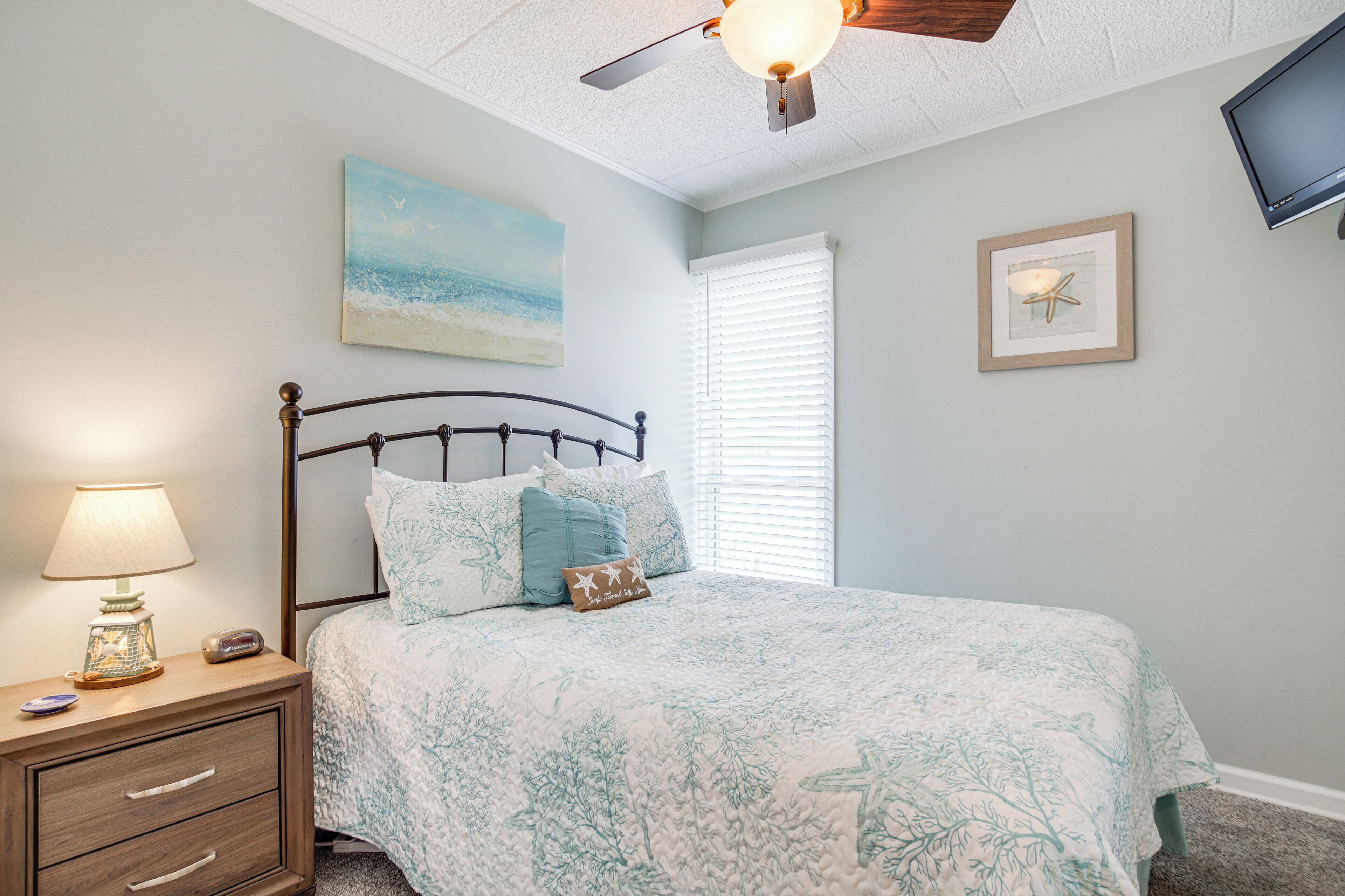 A bedroom with a bed, nightstand, lamp, ceiling fan, artwork, and a framed picture on the wall.