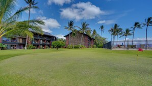 A well-maintained grassy area with a red flag marker, palm trees, and a building with a stone facade and multiple balconies. - Breezy Kaunakakai Retreat w/ Community Pool! (Kaunakakai)