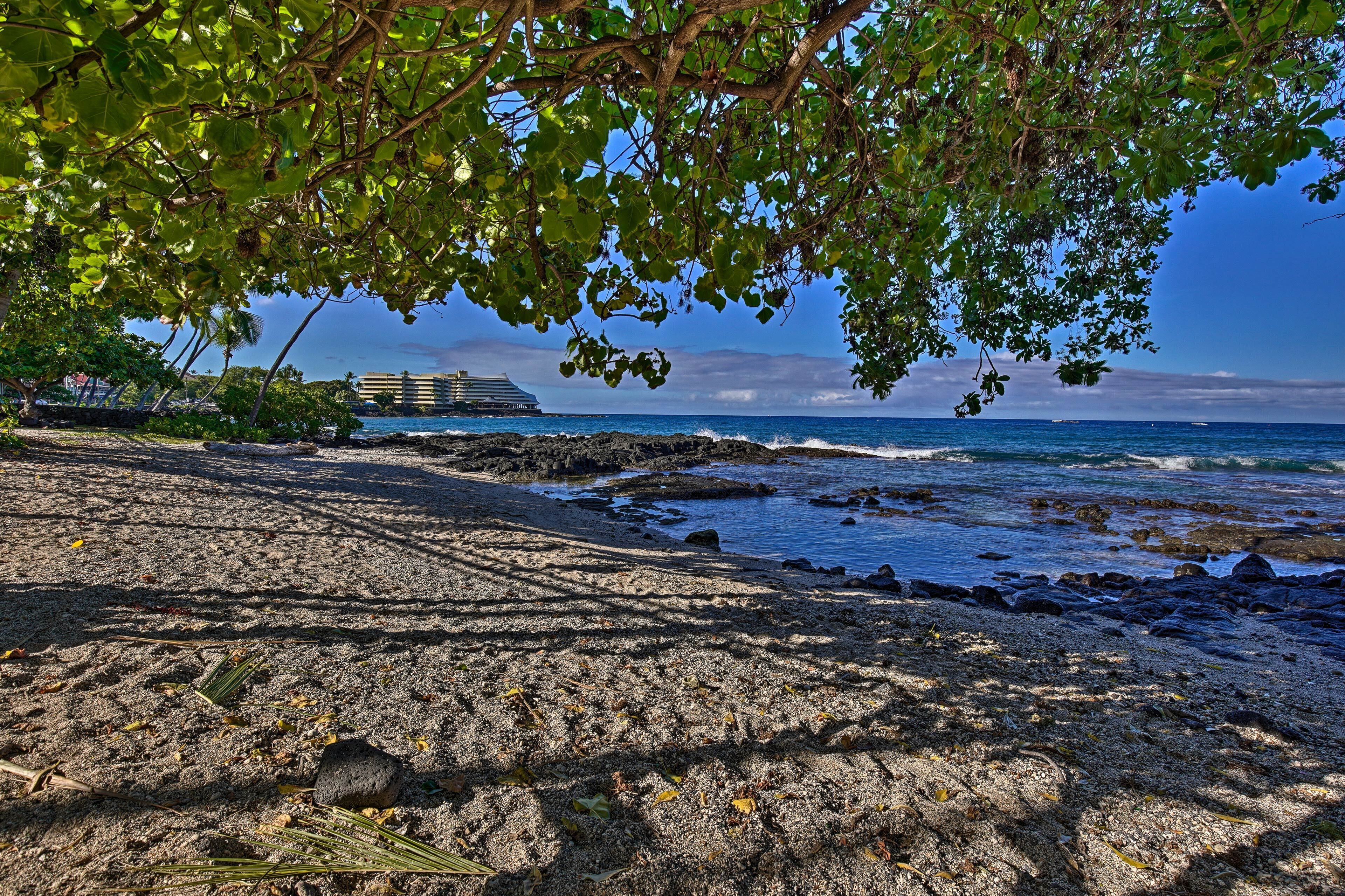 A beach with a rocky shoreline, a large cruise ship in the background, and a building with a sloped roof.