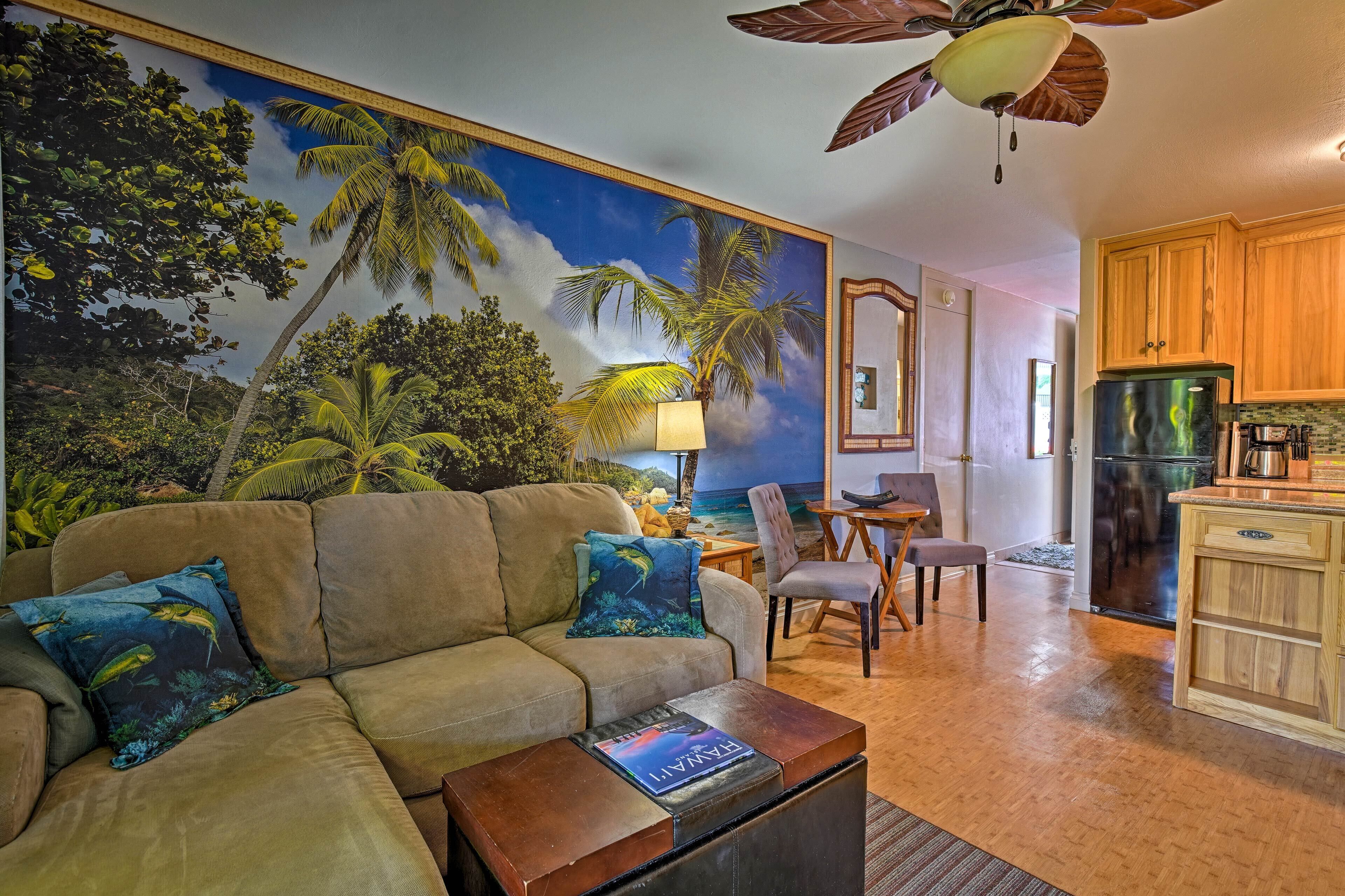 A living room with a large tropical mural, a sofa with decorative pillows, a coffee table with a magazine, a dining area with a table and chairs, and a kitchen with wooden cabinets and appliances.