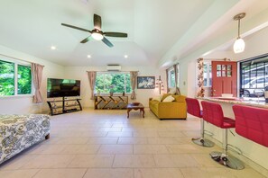 A spacious living room with a sofa, a TV, a ceiling fan, and a kitchen area with red bar stools. - Bright Haiku Studio: Patio + Rainforest View (Haiku)