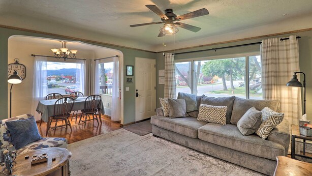 A living room with a sofa, a dining table, and chairs. There is a ceiling fan, a chandelier, and a window with curtains. - Comfy & Cozy Kalispell Home: Walk to Downtown (Kalispell)