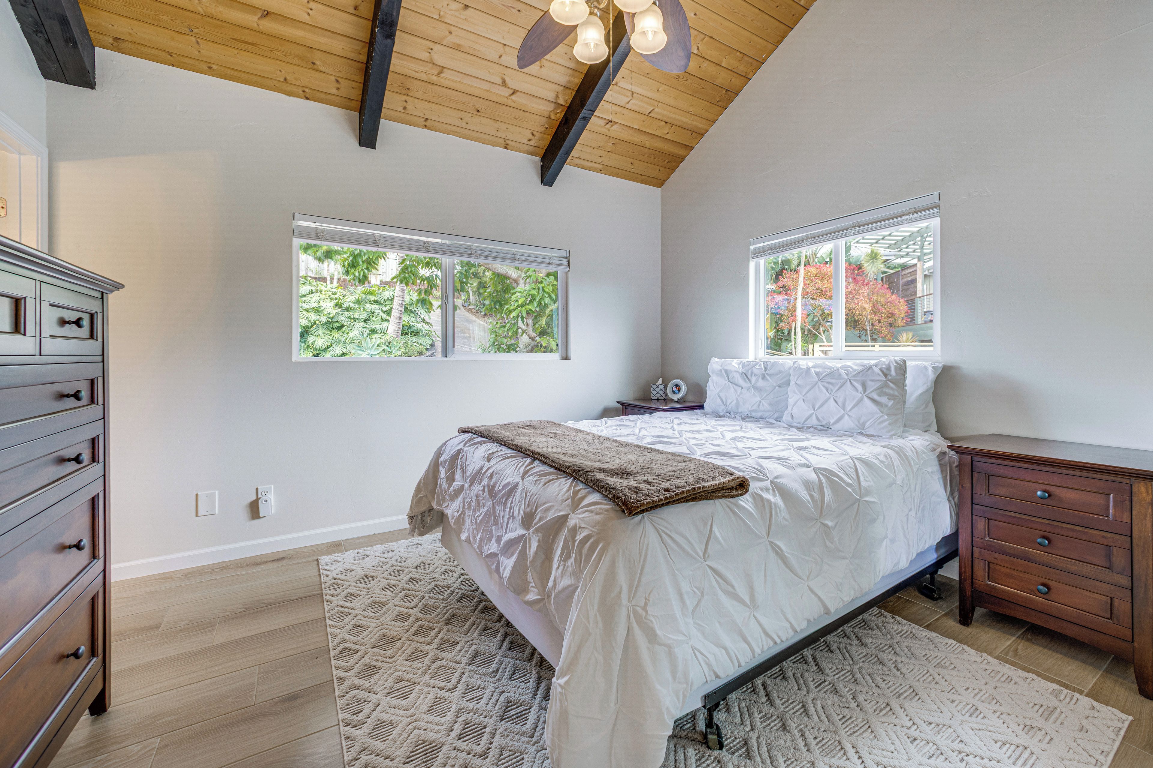 Un dormitorio con cama, mesita de noche, cómoda y dos ventanas.