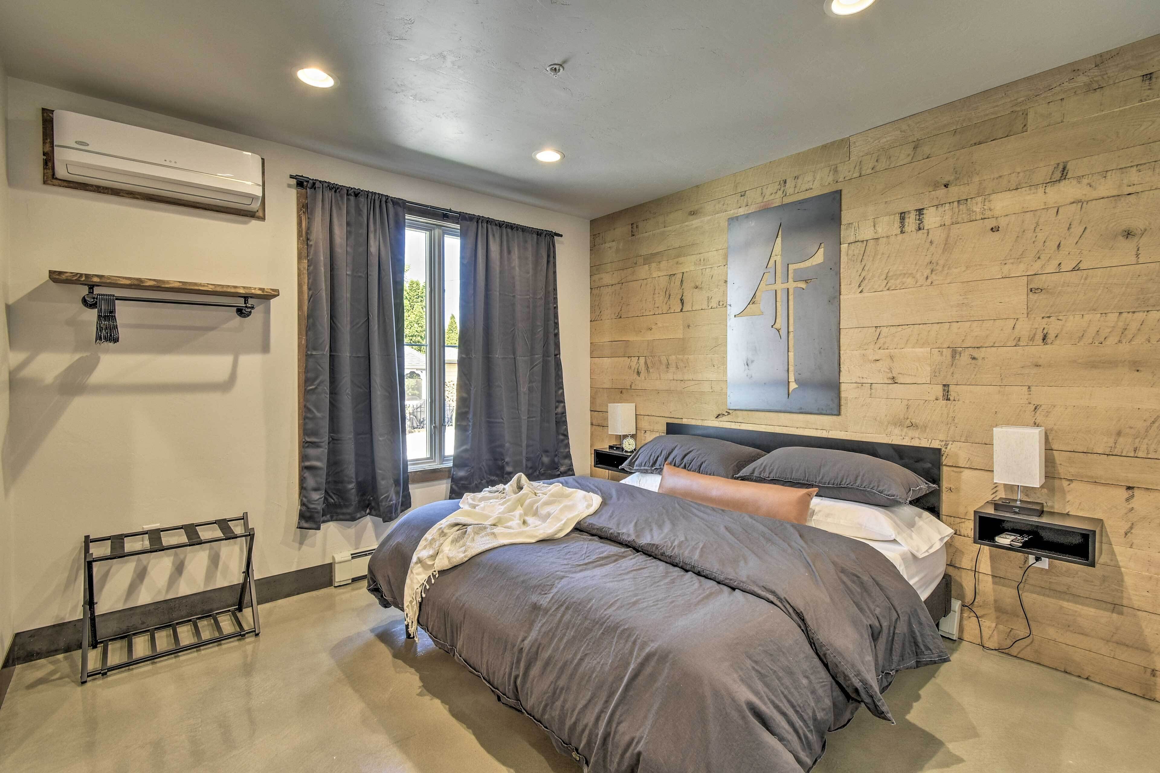 A modern bedroom with a large bed, a wooden wall panel, a mounted air conditioner, and a bedside table with a lamp.