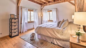 Un dormitorio con techo de madera, una cama grande, cortinas blancas, una mesita de noche con lámpara y un mueble de almacenamiento de mimbre.