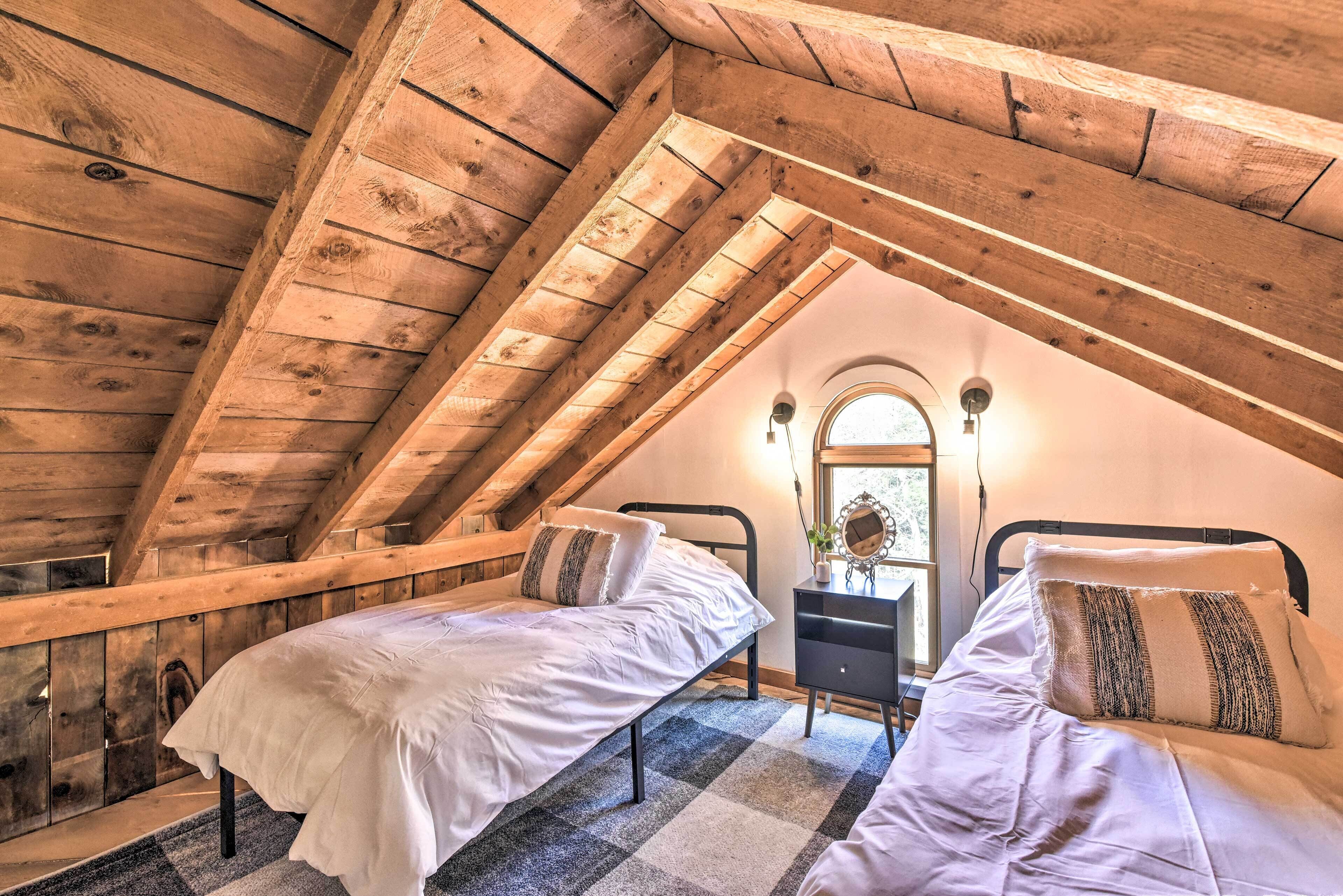 Une chambre mansardée chaleureuse avec deux lits, des poutres apparentes, une petite commode et une fenêtre offrant une belle vue.