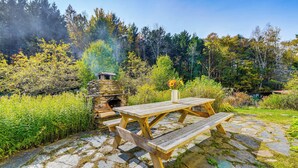 Un patio de piedra con una mesa de picnic de madera y bancos, rodeado de abundante vegetación y una chimenea de piedra que emite humo.