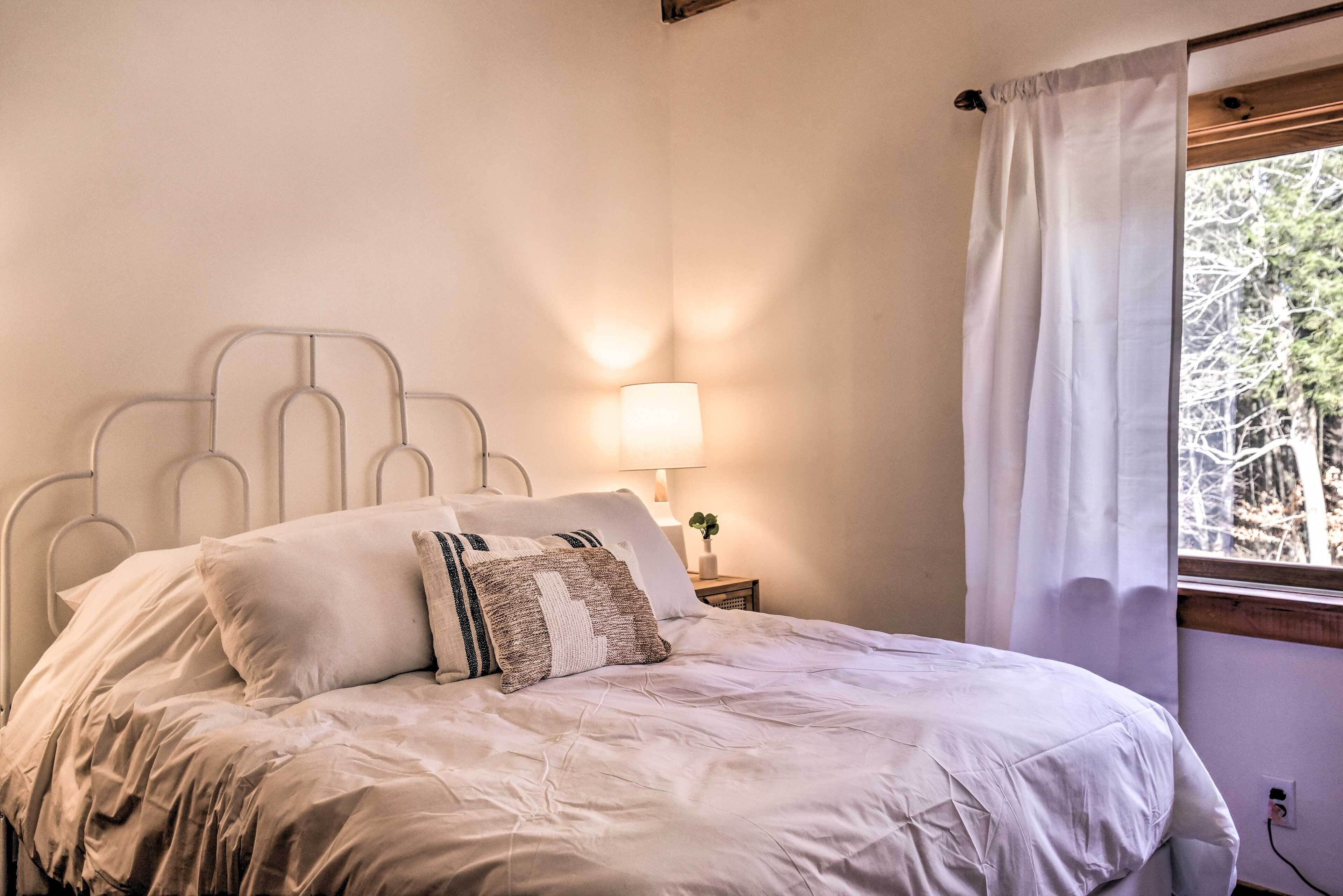 A neatly made bed with a headboard, a bedside lamp, and a window with curtains.