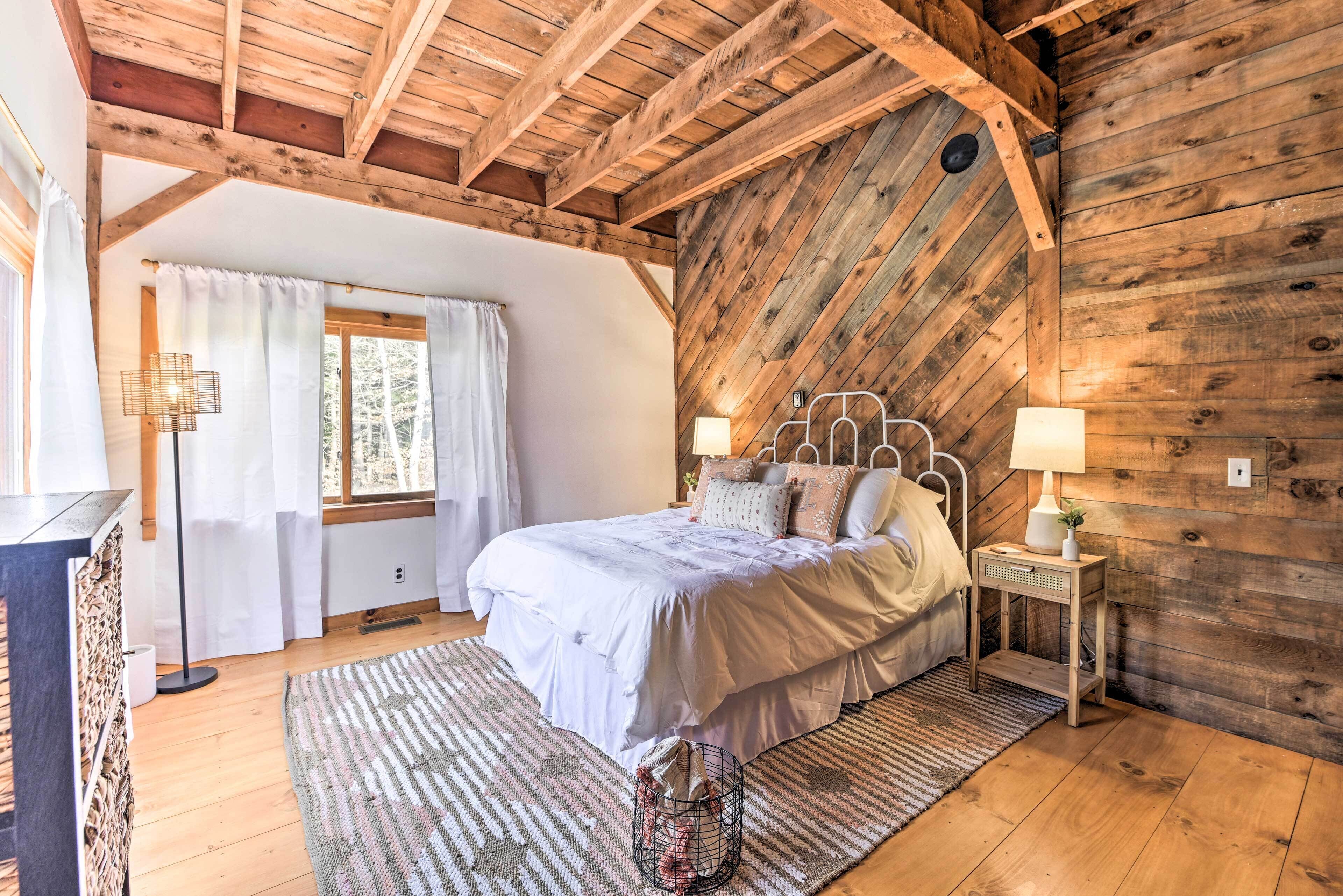 A bedroom with a wooden ceiling, a bed with white bedding, a nightstand with a lamp, and a window with curtains.
