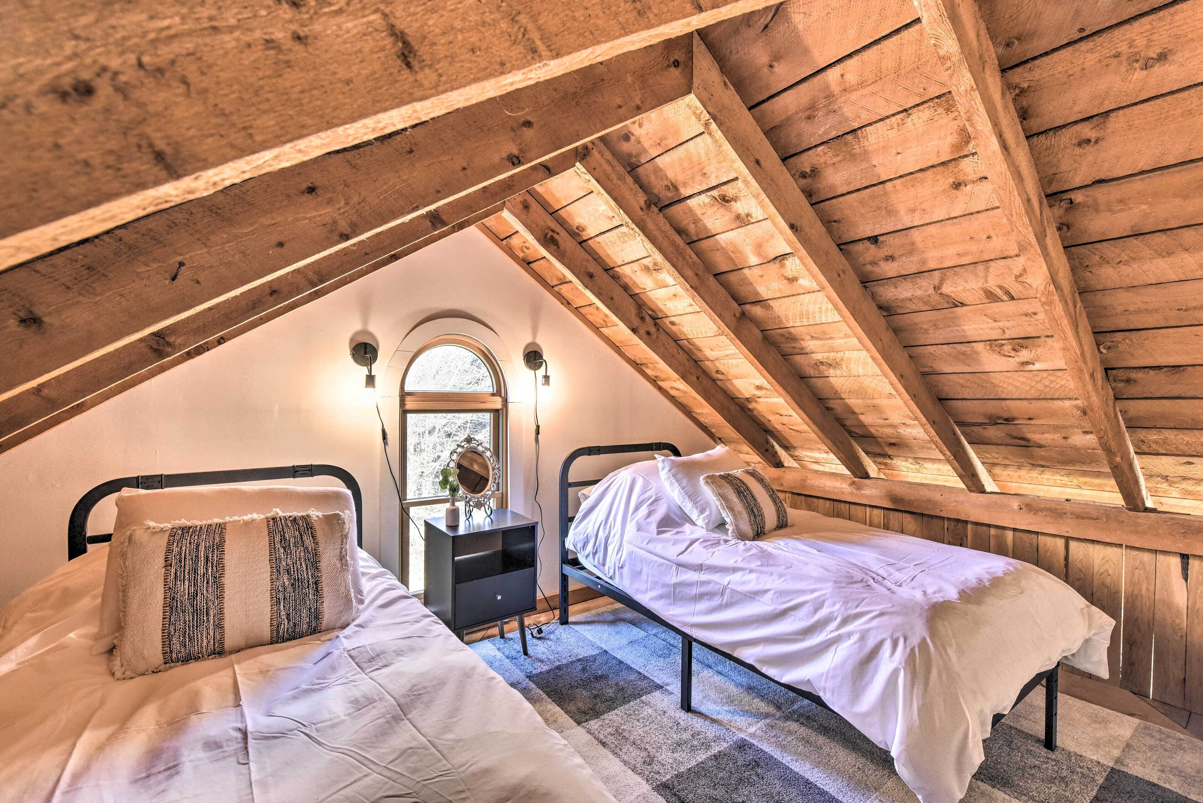 A cozy attic bedroom with two beds, a small dresser, and a window with a view of trees.
