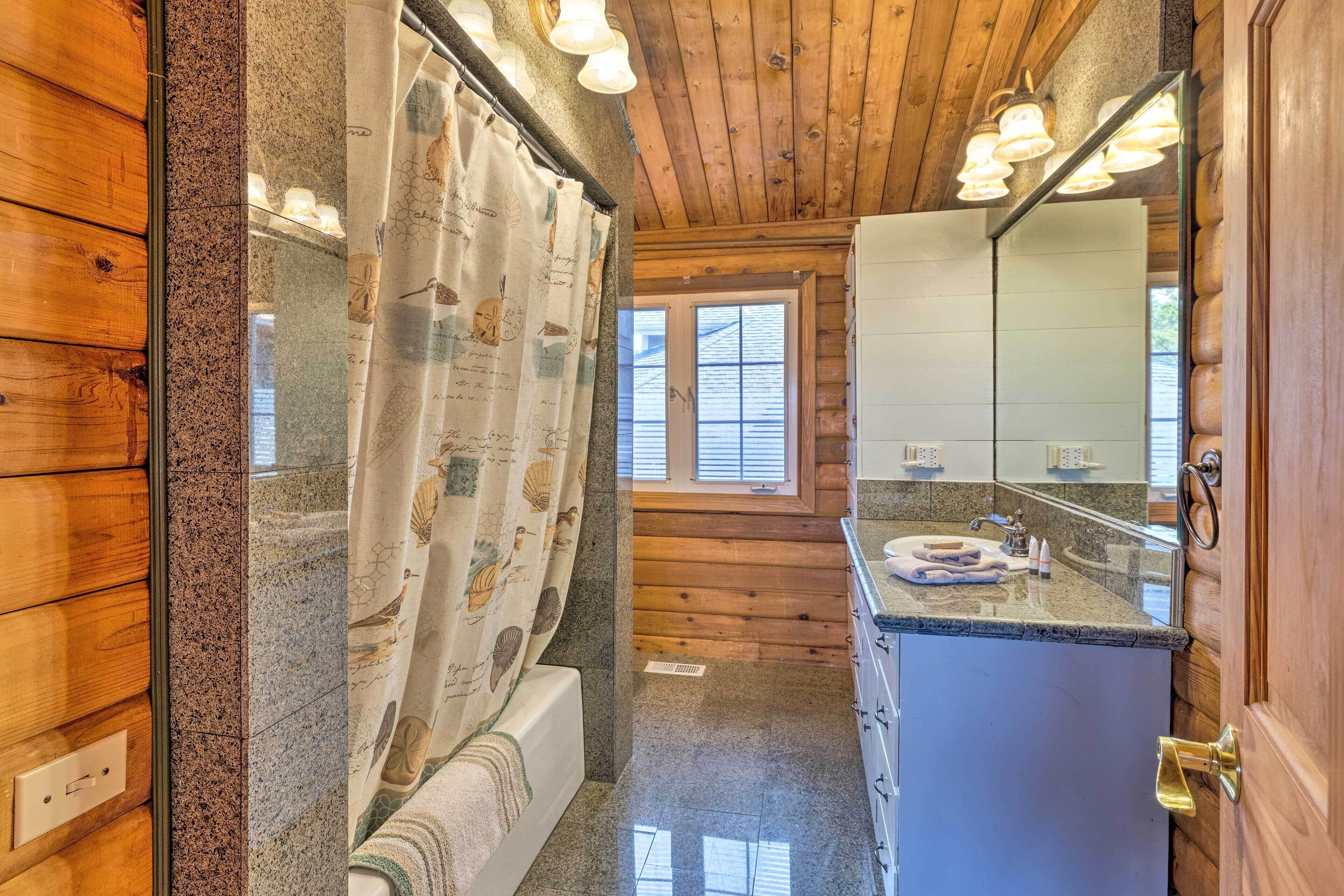 A bathroom with wooden walls, a shower curtain with a nature pattern, a granite countertop, and a mirror with lights above it.