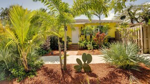 A yellow house with a front porch, surrounded by various tropical plants and a wooden fence. - West Palm Beach Home w/ Fenced-in Yard & Deck! (West Palm Beach)