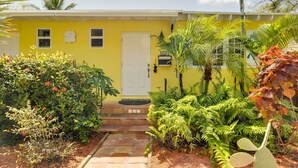 A yellow house with a white door, surrounded by lush greenery and a small garden. - West Palm Beach Home w/ Fenced-in Yard & Deck! (West Palm Beach)