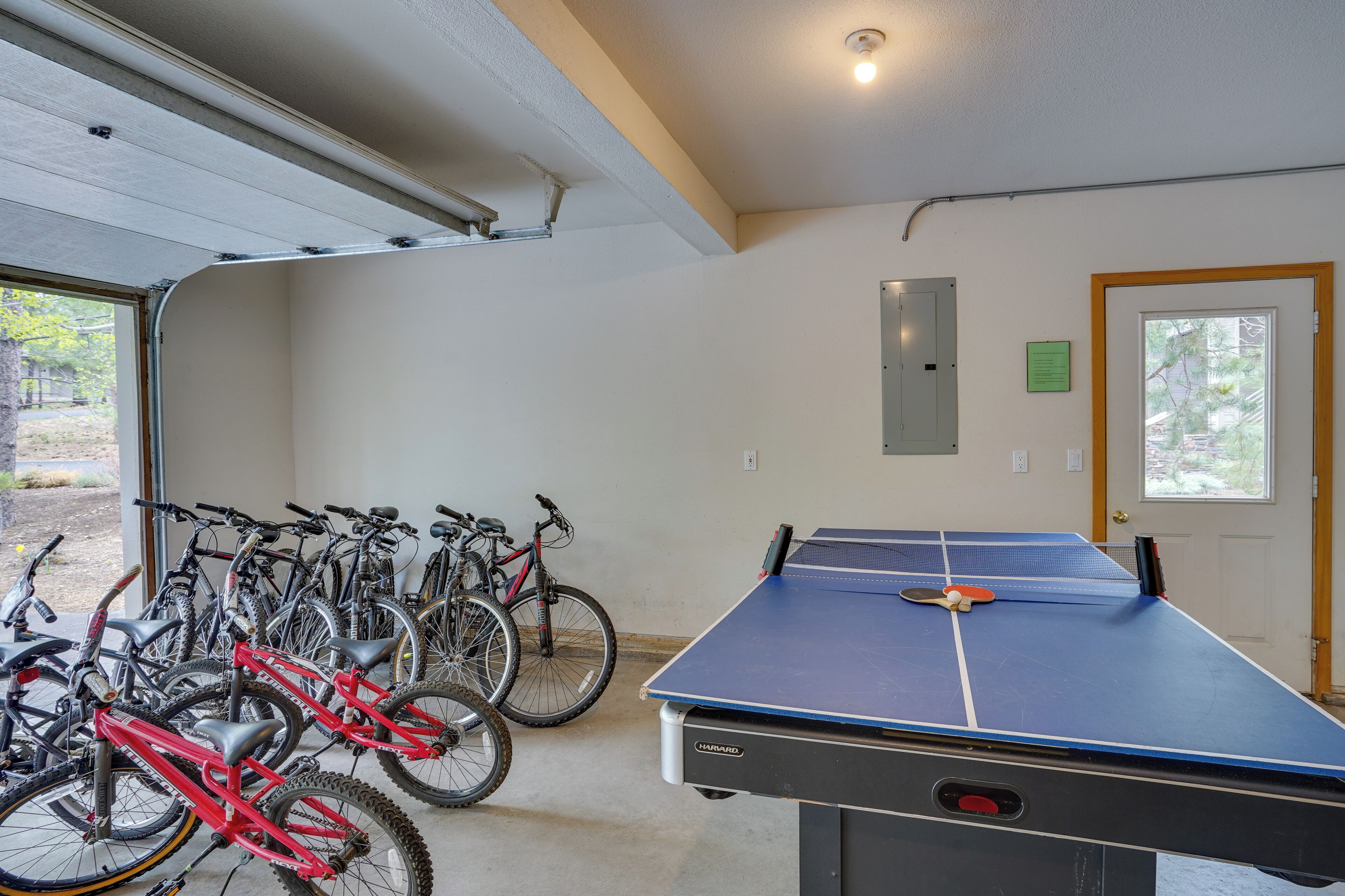 A room with a ping pong table, bicycles, and a door with a view of trees.