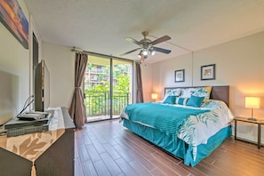 A bedroom with a bed, a ceiling fan, a TV, a desk, and a balcony with a view of greenery. - Kailua-kona Condo w/ 2 Private Lanais! (Kailua-Kona)