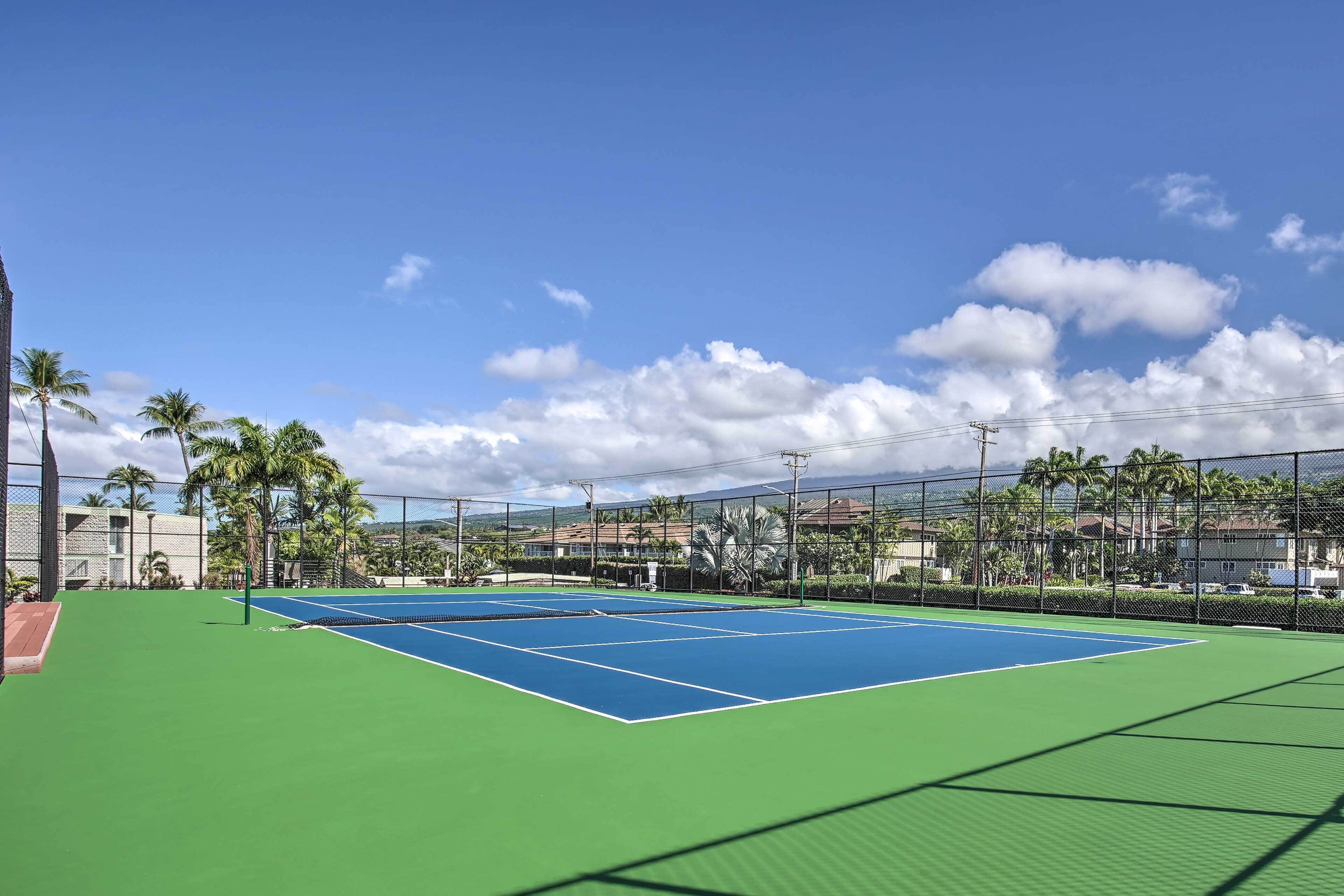 Una cancha de tenis exterior con superficie verde y líneas de demarcación azules, rodeada por una valla y palmeras.