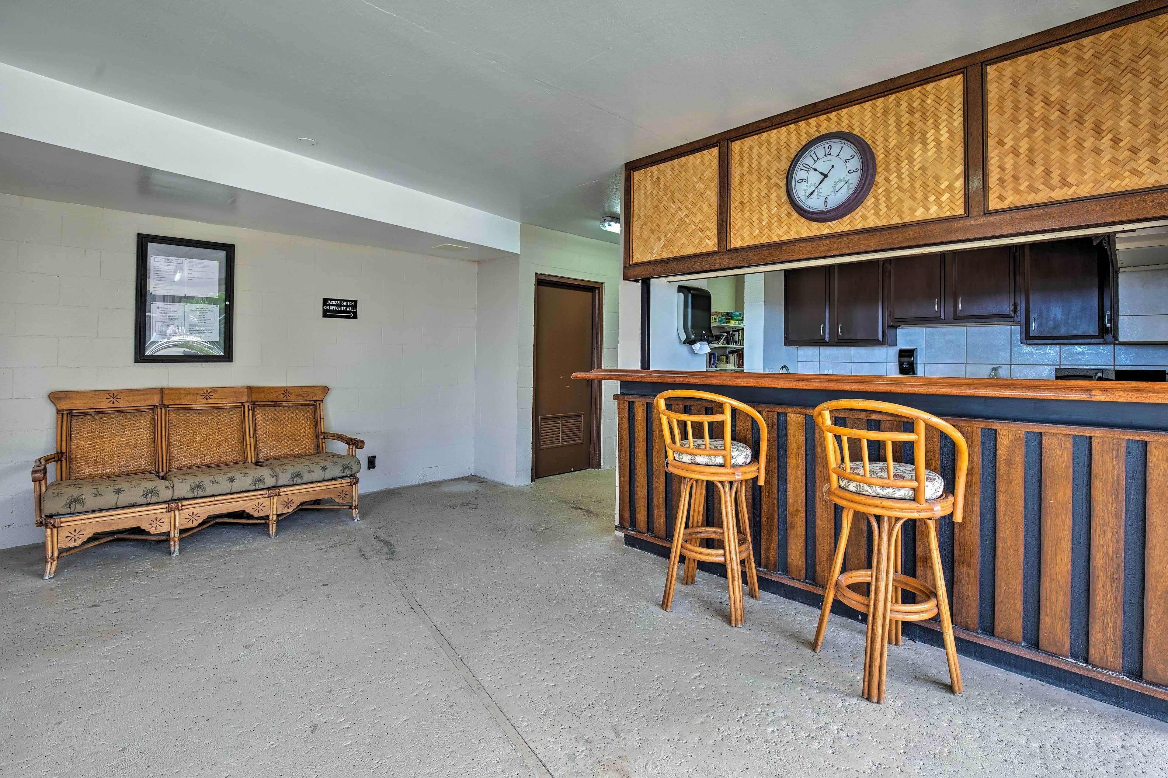 Une pièce comprenant un canapé en bois, deux tabourets de bar, un coin cuisine et une horloge au mur.