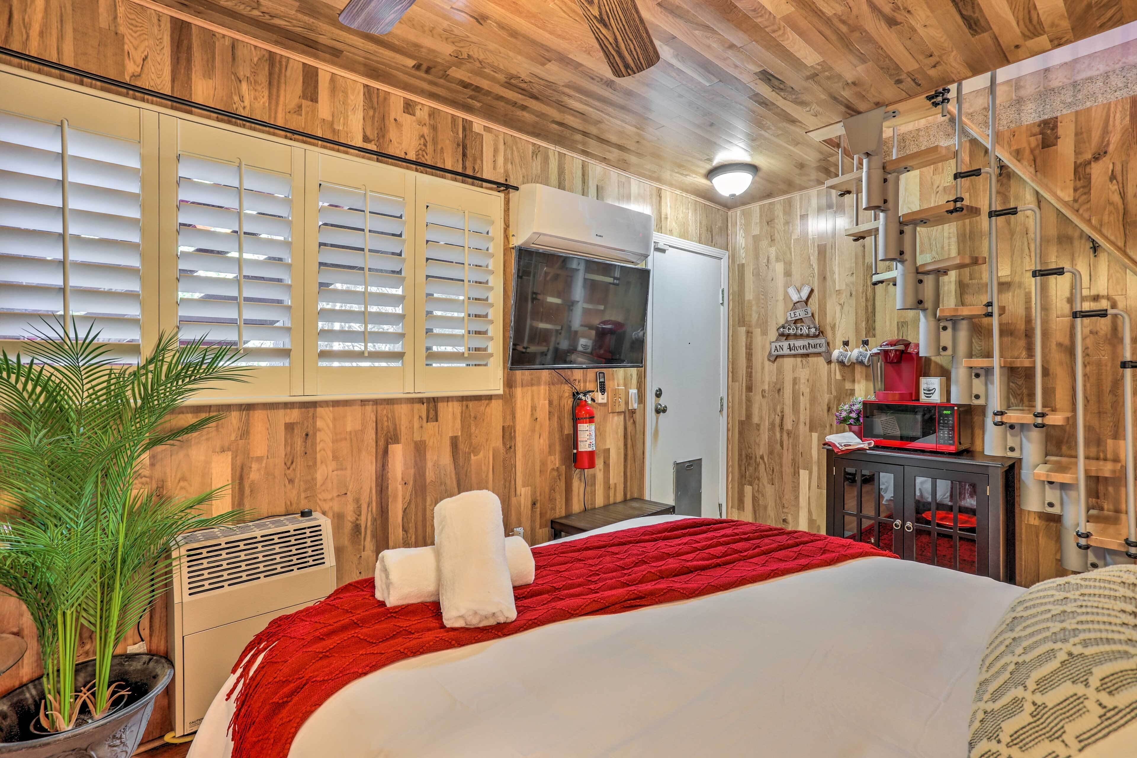 A hotel room with wooden walls, a bed with a red blanket, a potted plant, a small table, a microwave, and a fire extinguisher.