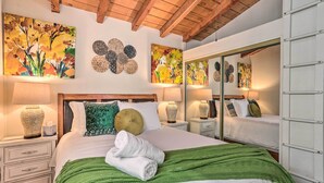 Une chambre bien éclairée avec un grand lit, un plaid vert et des coussins décoratifs. On y trouve également des œuvres d’art murales et un miroir au-dessus du lit.