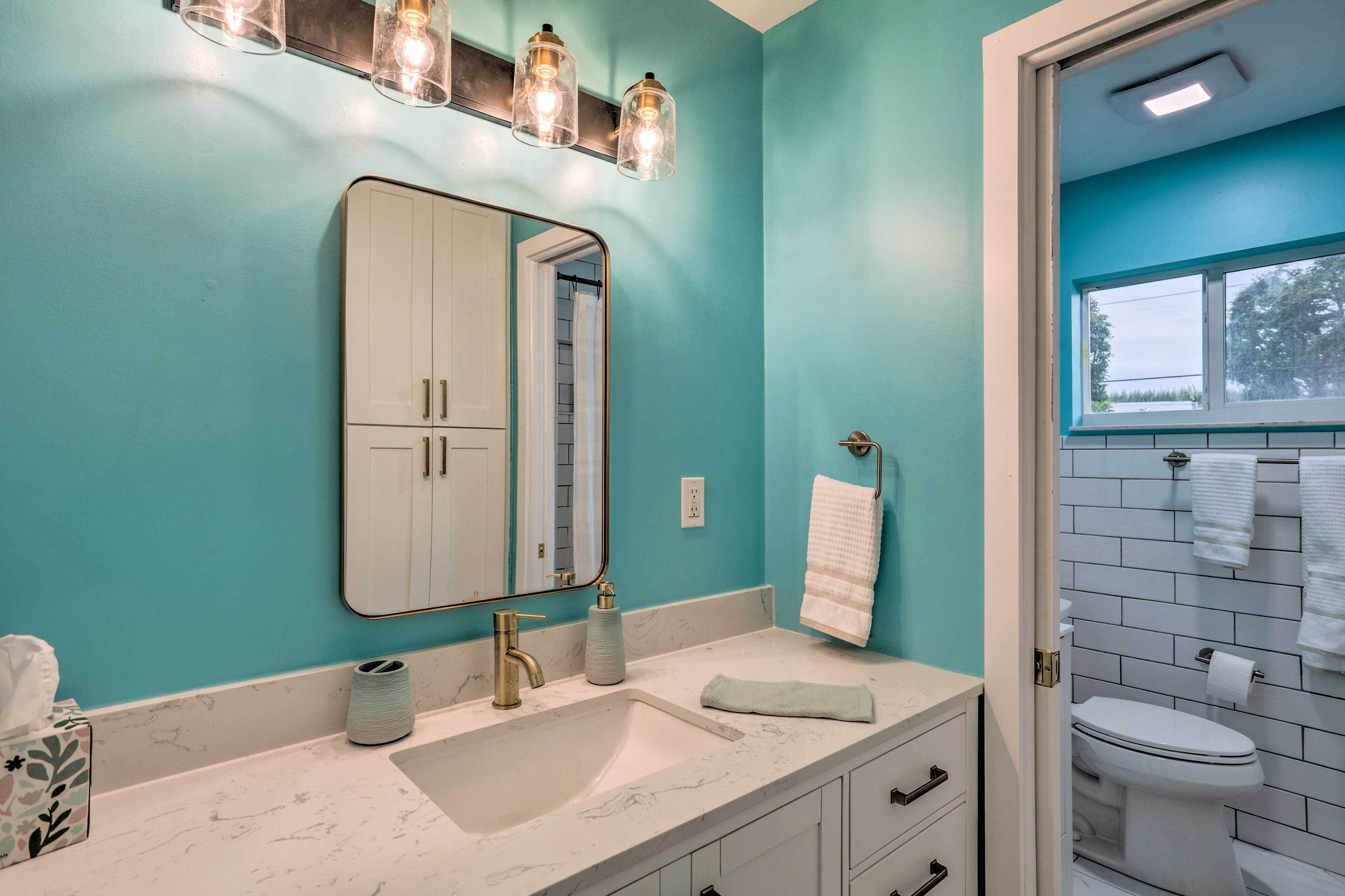 Casa de banho com parede azul-turquesa, lavatório branco com bancada de mármore, espelho grande e janela sobre o sanitário.