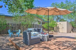 Une terrasse extérieure avec un canapé en osier, deux chaises bleues, une plateforme en bois et un grand parasol.
