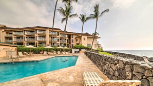A pool area with lounge chairs, a stone wall, and a building with balconies. - Oceanfront Kailua-kona Townhome w/ Pool & Views! (Kailua-Kona)