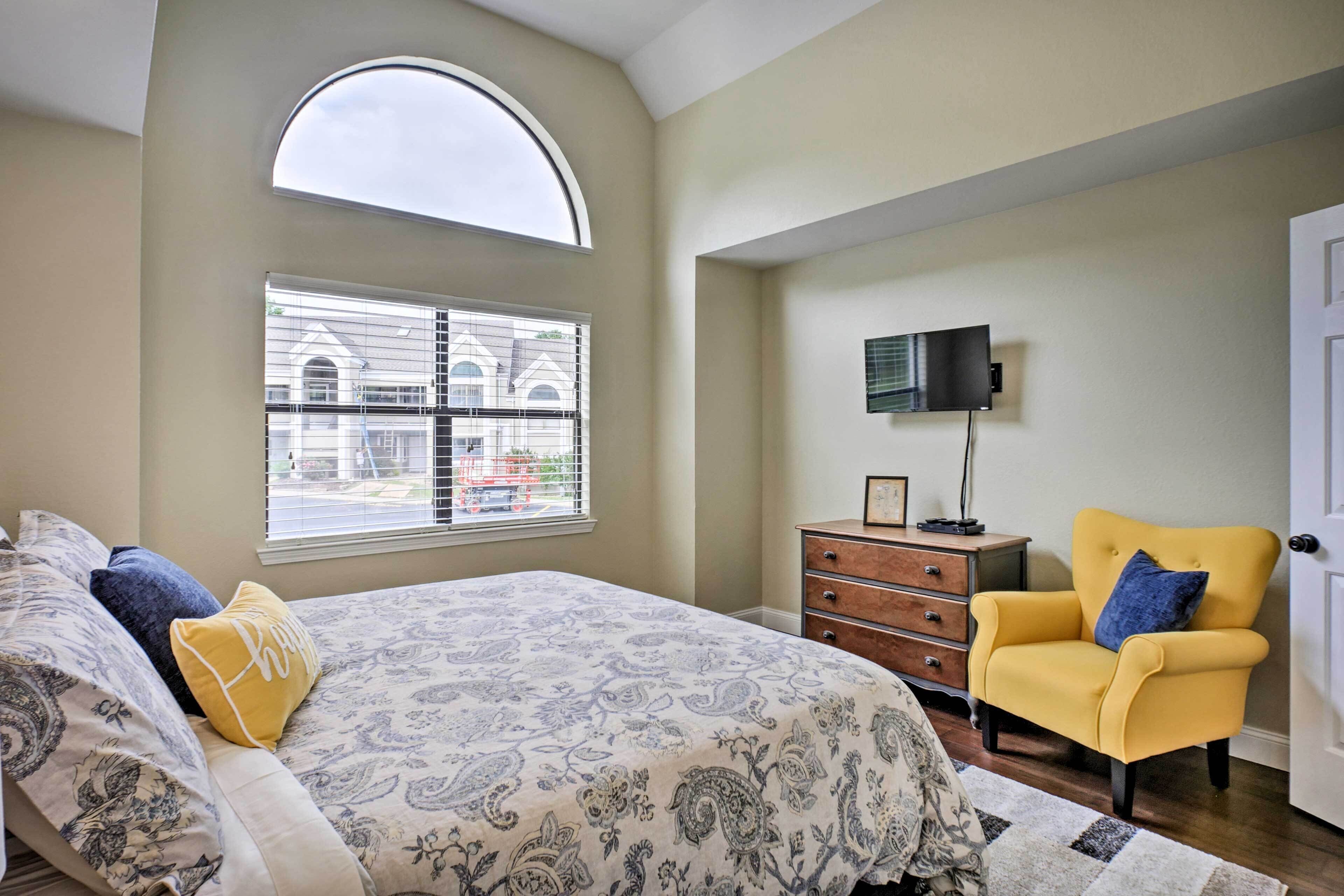Un dormitorio con cama, una silla, cómoda, televisor y ventana con persianas.