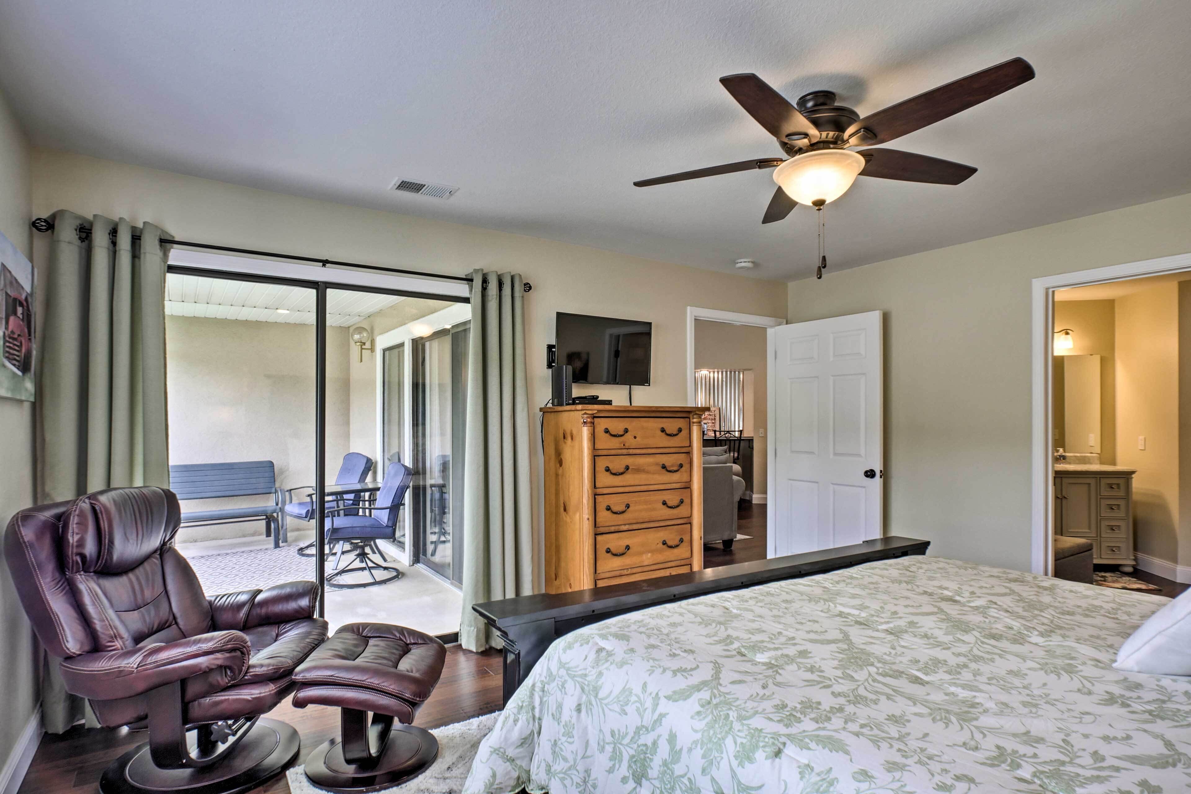Une chambre à coucher comprenant un lit, un fauteuil massant, une commode en bois, un ventilateur de plafond et une porte-fenêtre coulissante donnant sur un balcon.