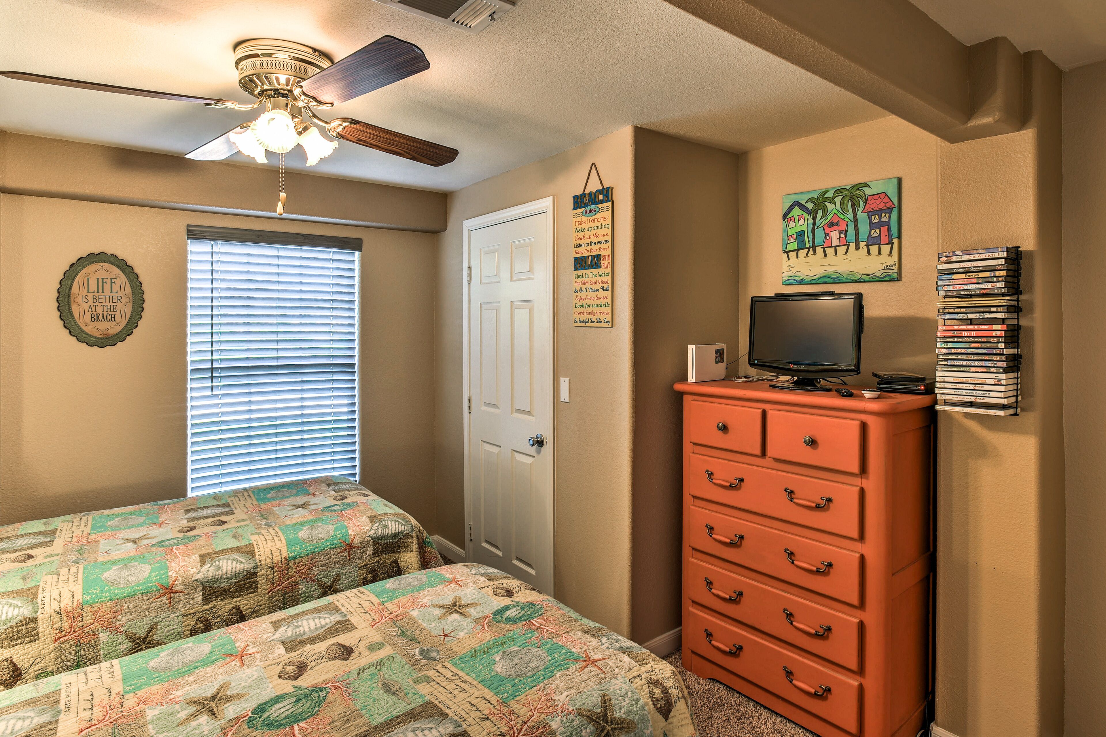 A bedroom with two beds, a ceiling fan, a dresser, a TV, and a wall with a beach-themed painting and a calendar.
