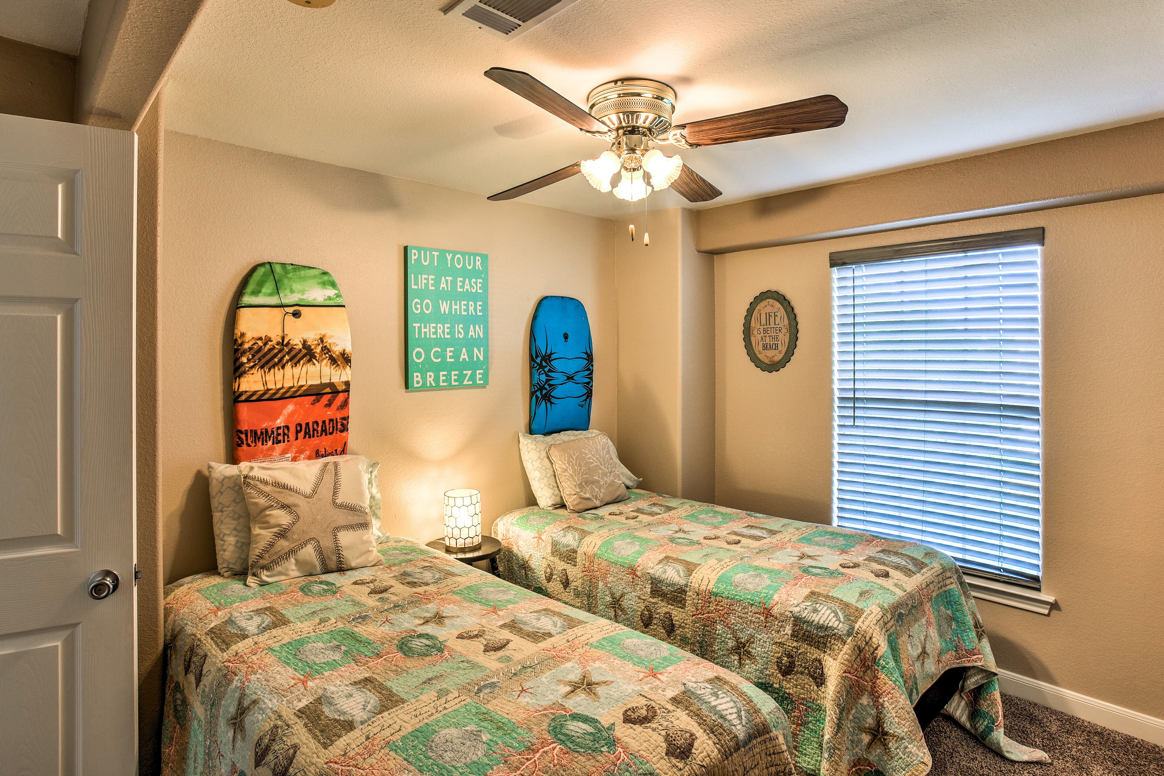 A bedroom with two beds, a ceiling fan, a window with blinds, and wall decorations including surfboards and a motivational quote.