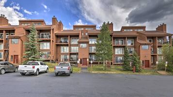 Un edificio residencial de varios pisos con un diseño arquitectónico moderno, con balcones, ventanas amplias y un área de estacionamiento con varios vehículos.