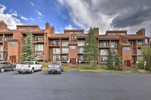 A multi-story residential building with a modern architectural design, featuring balconies, large windows, and a parking area with several vehicles.