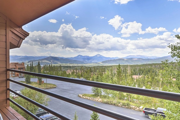 A balcony with a view of a mountain range, a parking area, and a building with a red roof.