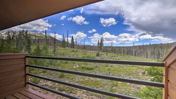 Una terraza de madera con barandillas de metal que da a un paisaje montañoso con árboles dispersos y cielo despejado.