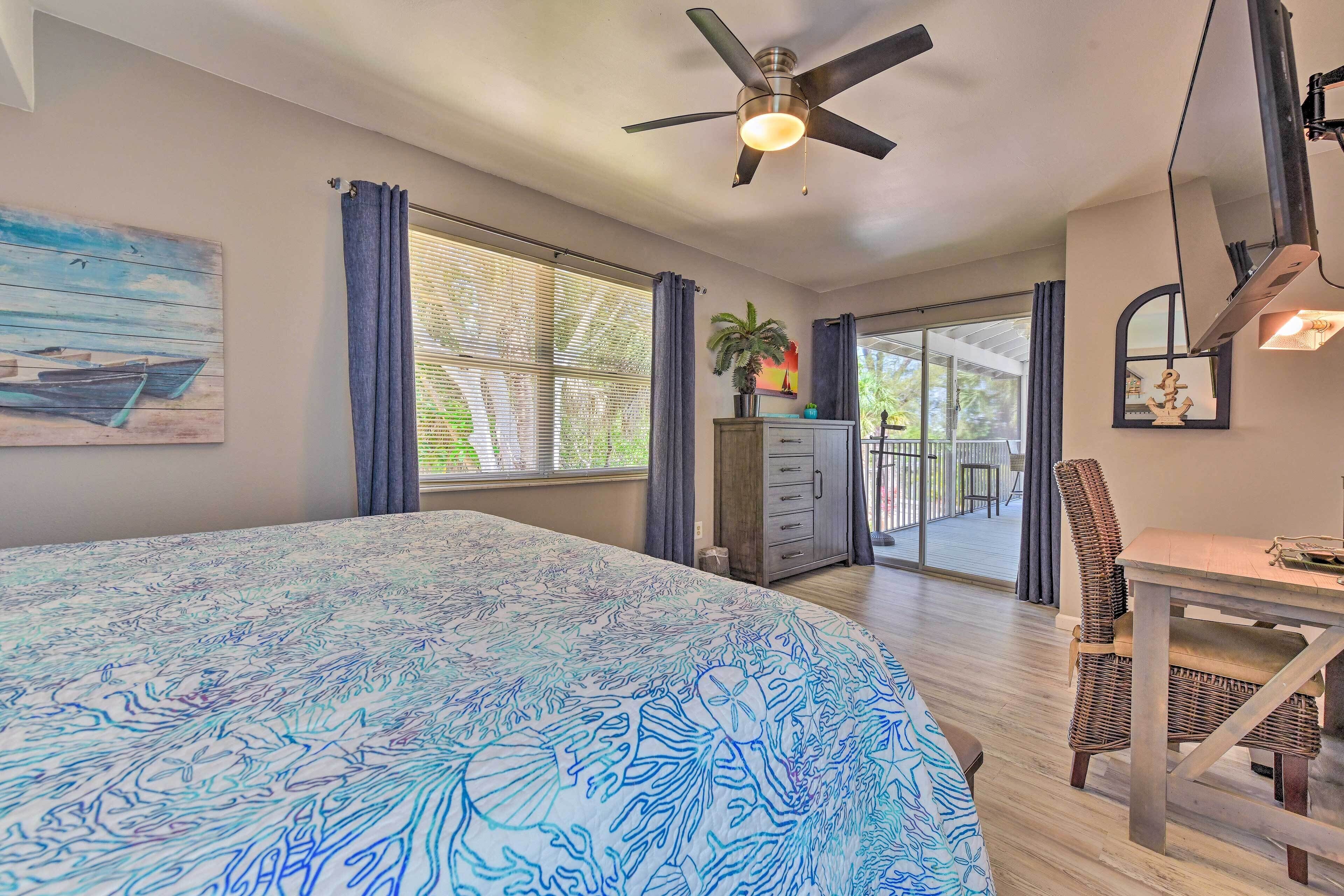 A bedroom with a bed, a ceiling fan, a window with curtains, a desk with a chair, and a balcony.