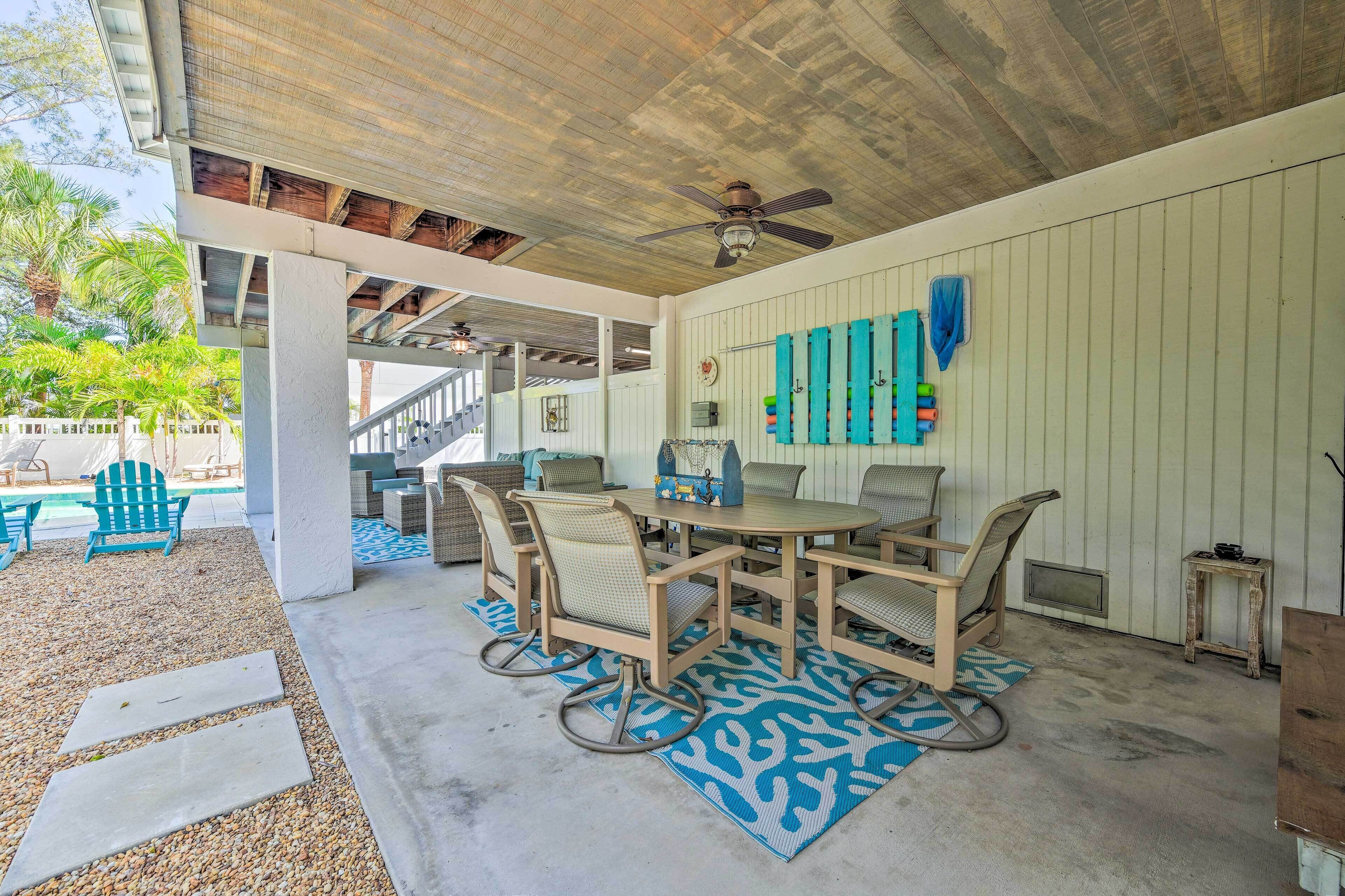 A covered outdoor patio with a round table and chairs, a ceiling fan, and a view of a pool and palm trees.