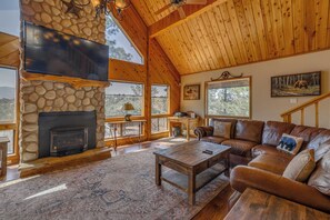 A spacious living room with a stone fireplace, leather sofa, and large windows offering a view of the outdoors. - Tranquil Ruidoso Cabin w/ Private Hot Tub! (Ruidoso)