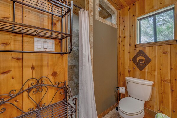 A wooden cabin bathroom with a shower, a toilet, a metal bench, and a wall-mounted light switch. - Tranquil Ruidoso Cabin w/ Private Hot Tub! (Ruidoso)