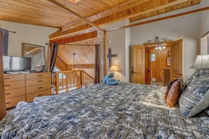 A bedroom with a bed, wooden ceiling, a desk with a computer, and a door leading to another room.