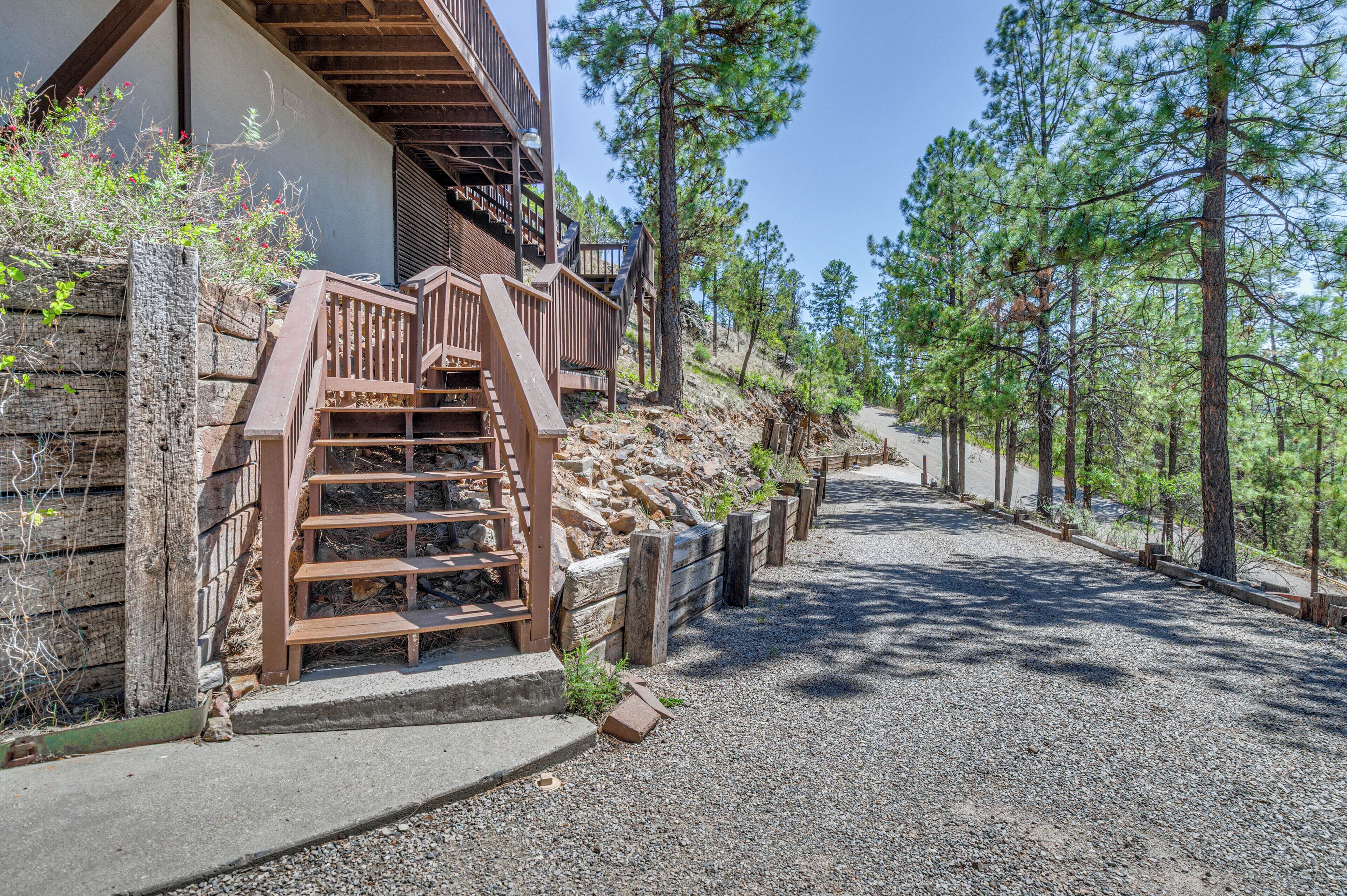A building with a wooden staircase leading to a deck, surrounded by trees and a gravel path.