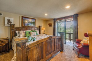 A bedroom with a wooden bed, a nightstand, a lamp, a chair, and a balcony with a view. - Tranquil Ruidoso Cabin w/ Private Hot Tub! (Ruidoso)