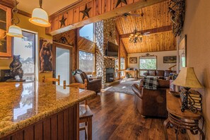 A spacious living area with a stone fireplace, wooden ceiling beams, and a large window. - Tranquil Ruidoso Cabin w/ Private Hot Tub! (Ruidoso)