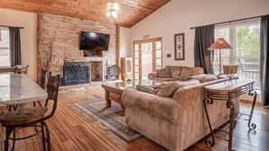 A spacious living room with a stone fireplace, a large flat-screen TV, a sofa, and a dining area with a table and chairs. - Stunning Ruidoso Cabin w/ Private Hot Tub! (Ruidoso)