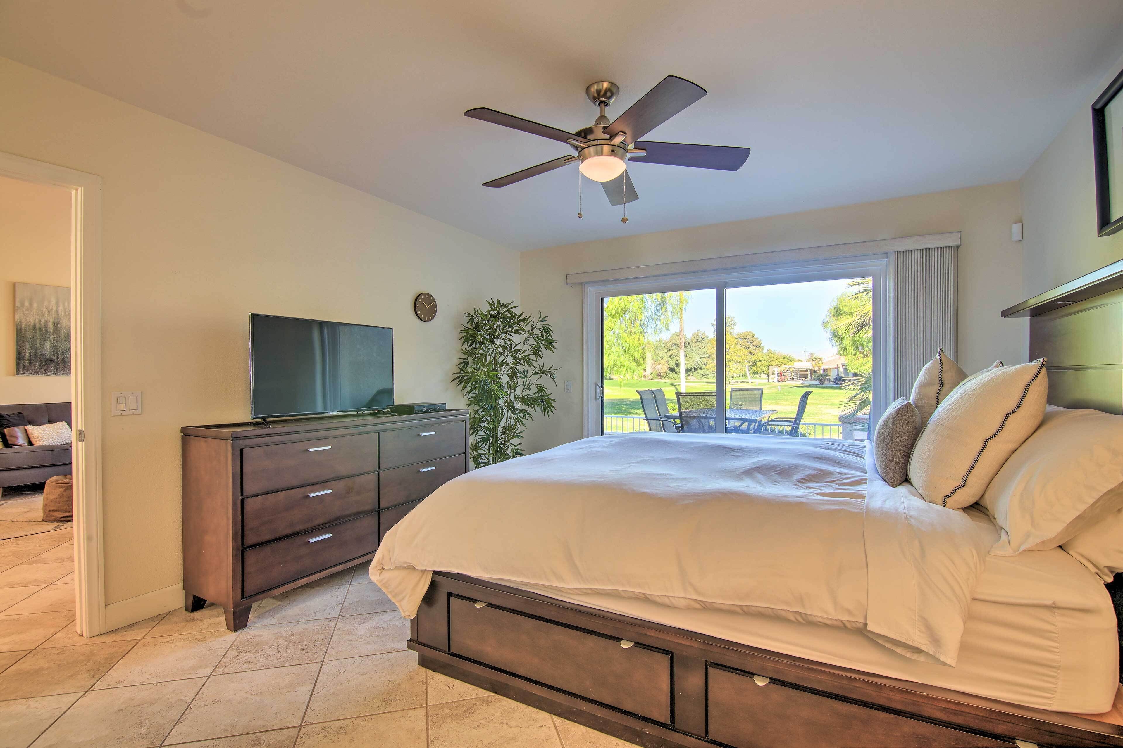 A bedroom with a bed, a dresser, a television, a ceiling fan, and a large window with a view of the outdoors.