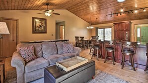 A spacious living room with a wooden ceiling, a grey sofa with patterned cushions, a coffee table, and a dining area with wooden chairs and a table. - Cabin w/ Fire Pit & Movie Room, 1 Mi to Lake (Pequot Lakes)