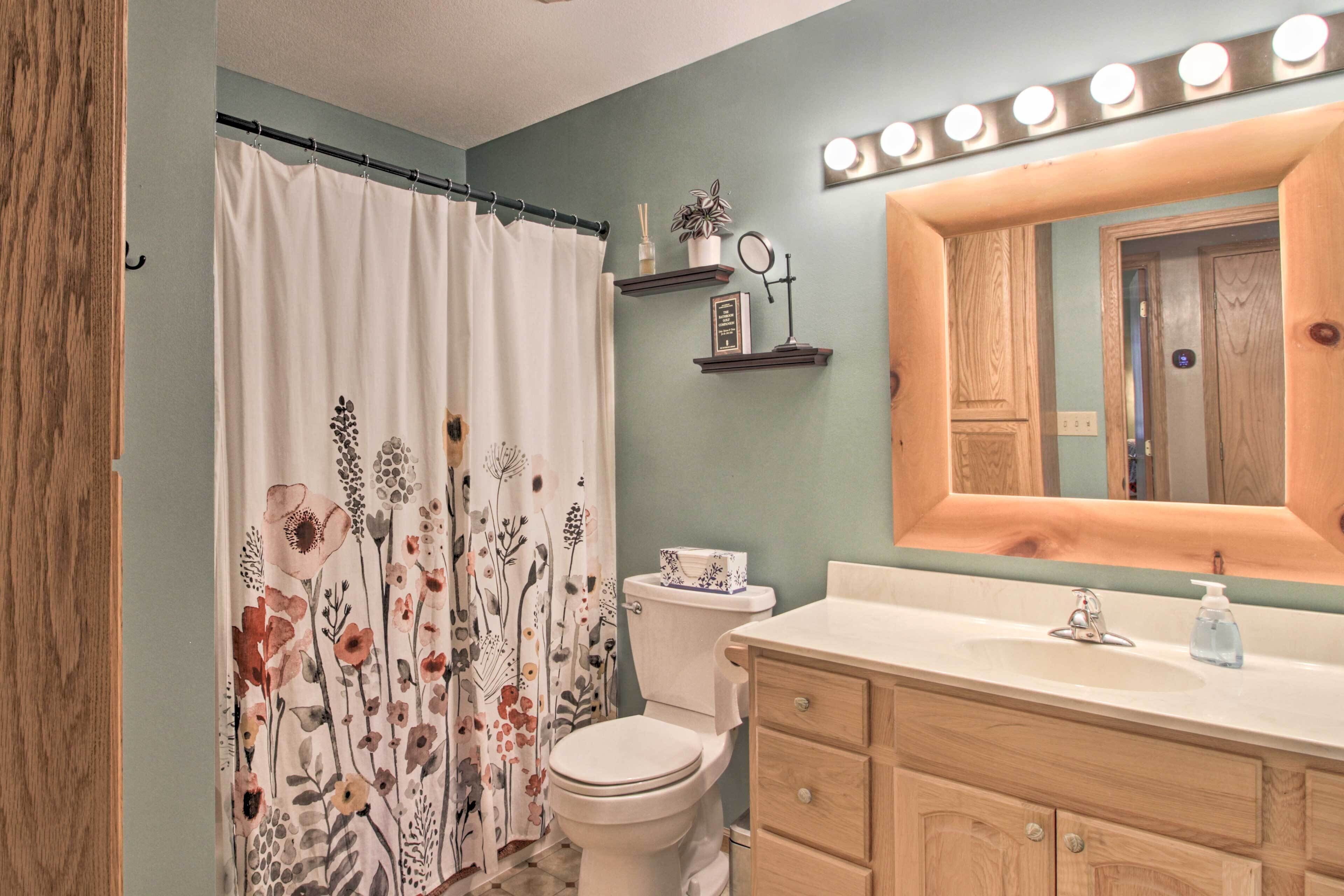 Un baño con cortina de ducha estampada con motivos florales, un inodoro, un tocador con espejo y un estante con artículos de aseo.